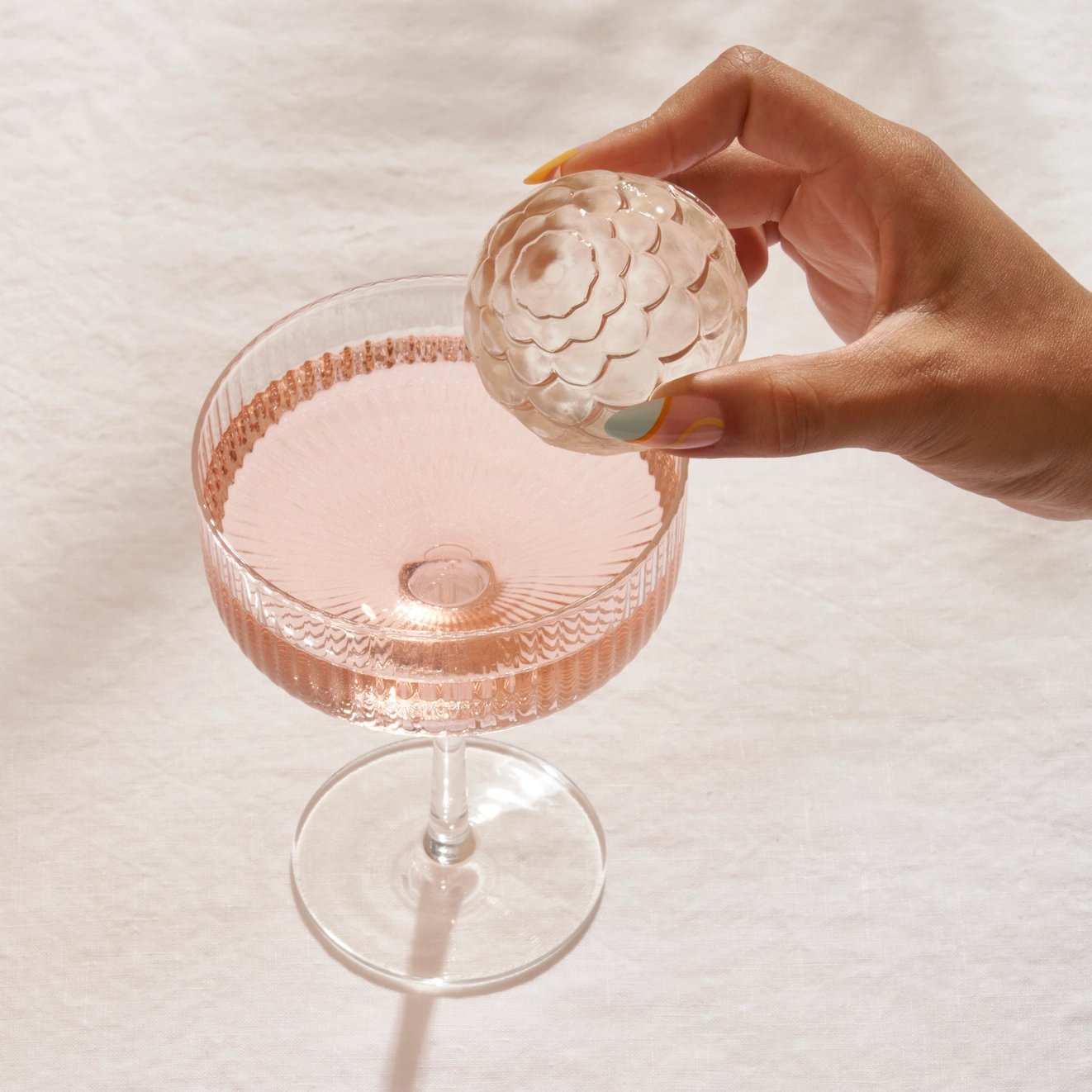 A person's hand gently placing an intricate ice cube into an elegant coupe glass filled with a pink beverage, set against a neutral textured backdrop.