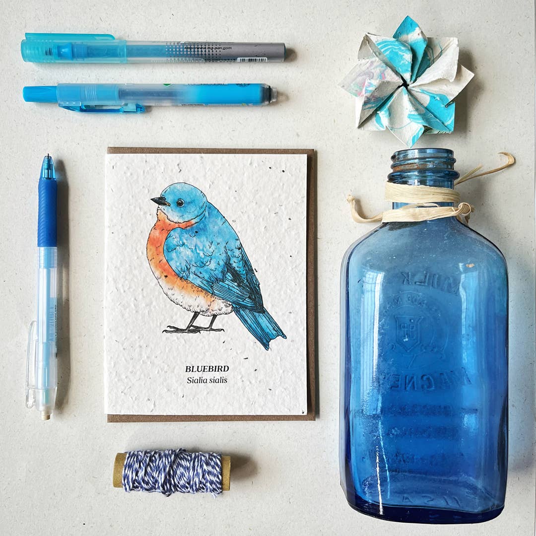 A neatly arranged assortment of art supplies including blue pens and threads with a Small Victories Bluebird Plantable Wildflower Seed Card next to a vintage blue glass bottle, all laid out on a clean, light surface, capturing
